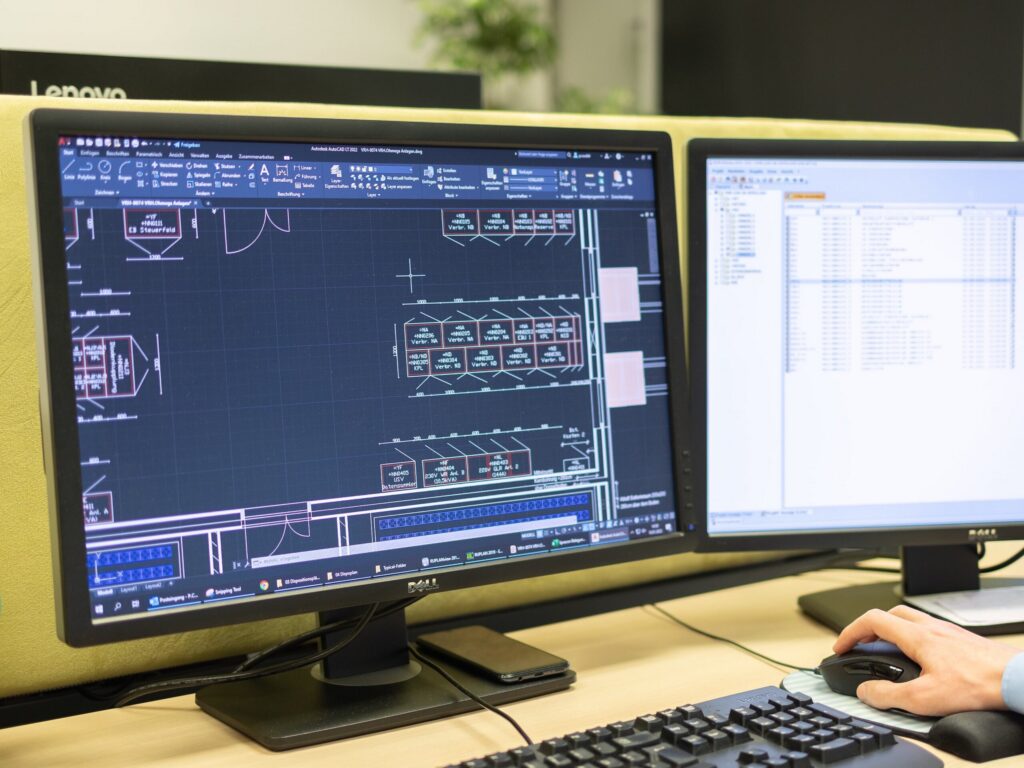 Zwei Monitore mit CAD-Plan einer Schaltanlagen-Aufstellung und Projektliste, Hand an der Maus