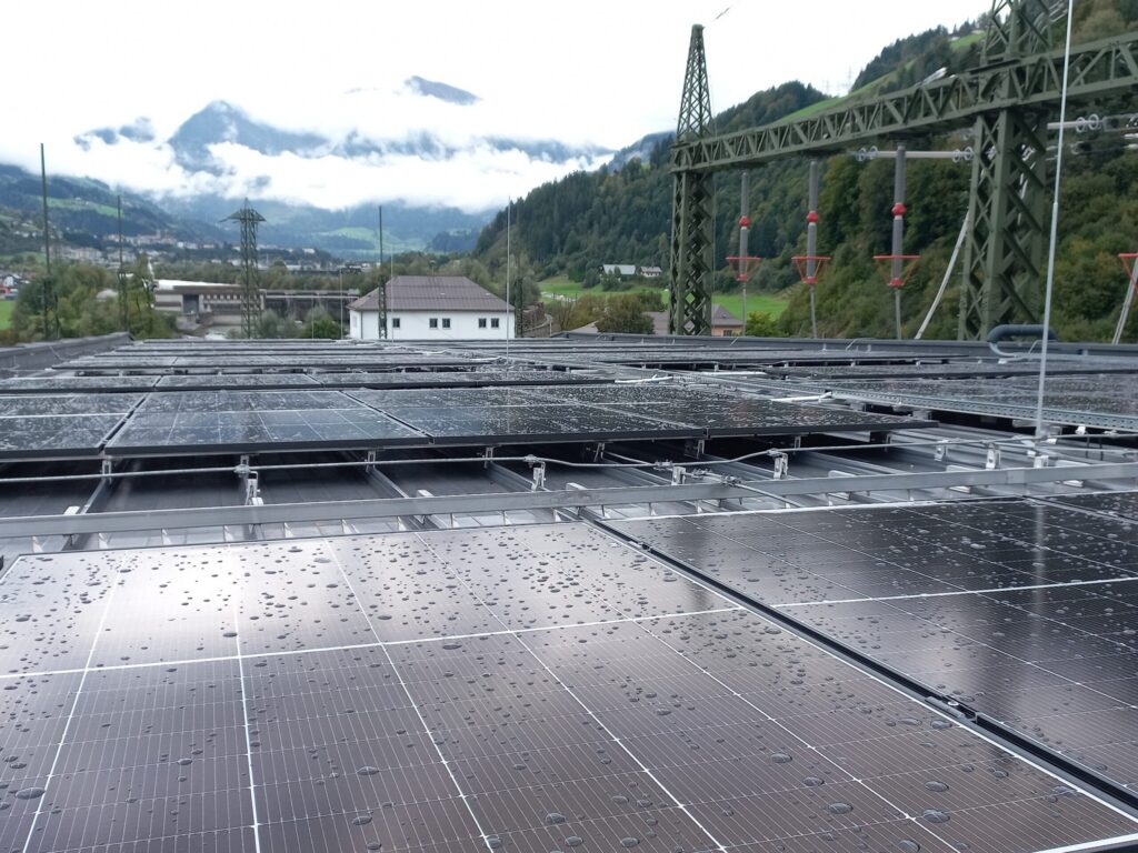 Regennasse Photovoltaikmodule auf einem Dach, im Hintergrund ein Umspannwerk und Berge