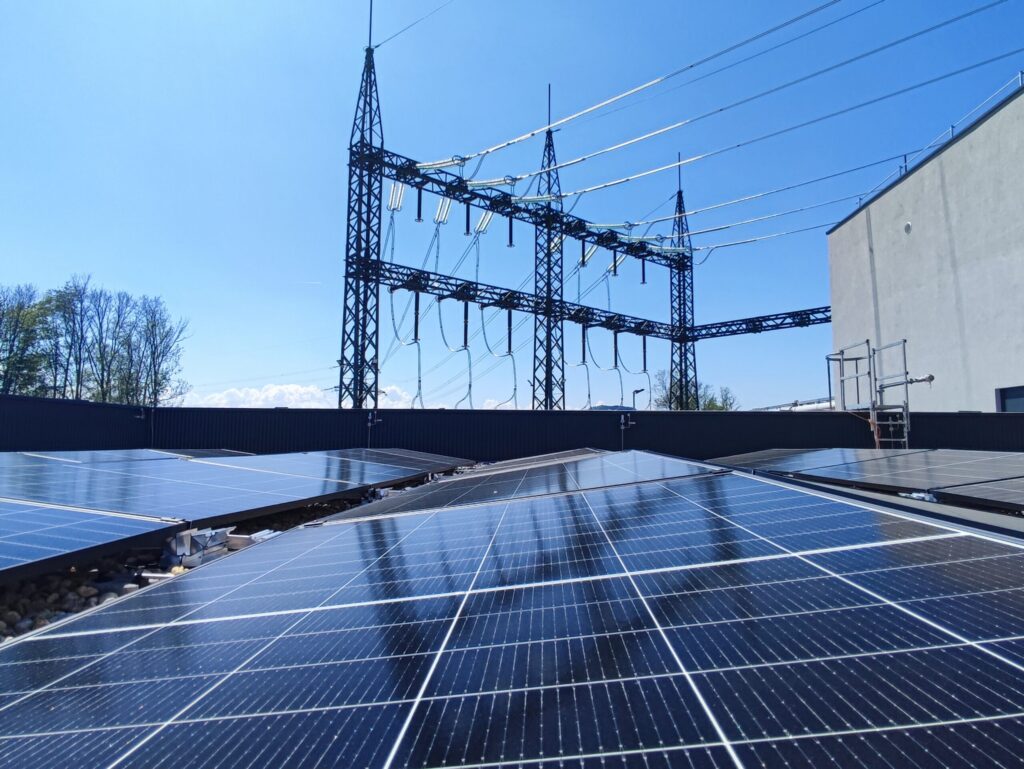 Photovoltaikmodule auf einem Dach vor einem Umspannwerk mit Freileitungen unter blauem Himmel